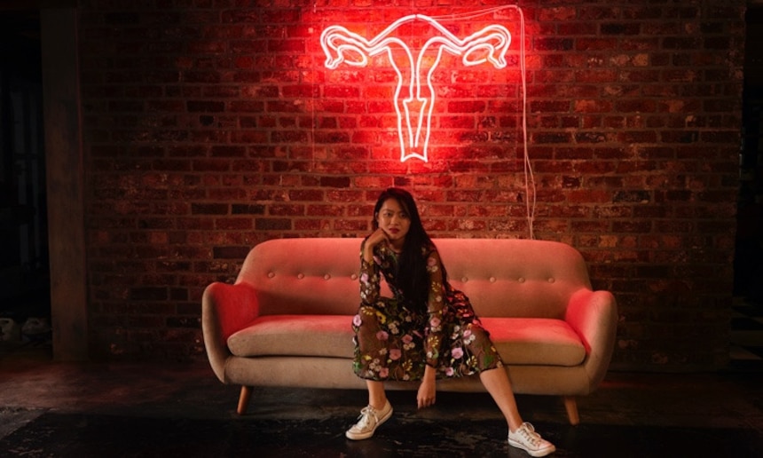 Kvinde i blomstret kjole, som sidder på en sofa foran en livmoder i neon