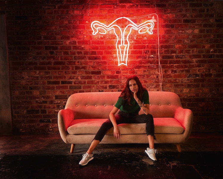 Woman sitting on a couch in front of a neon sigh in the shape of the female reproductive system | Bodyform