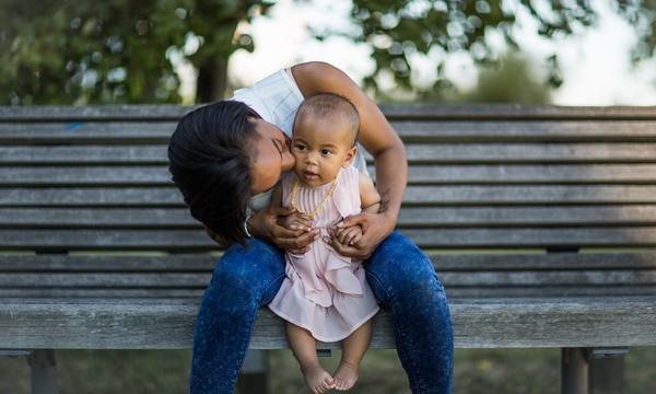 New mum kissing baby while sat on bench. Baby wearing pink dress. New mum in jeans and white top.