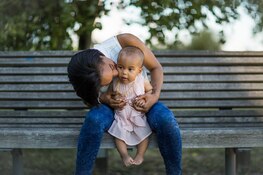 Nouvelle maman assise sur un banc embrassant son bébé. Bébé portant une robe rose. Nouvelle maman en jean et haut blanc.